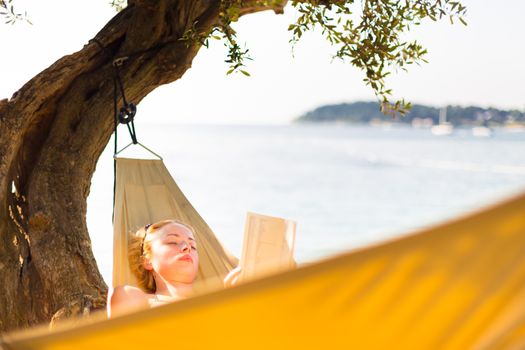 Beautiful girl lies on hammock on the beach reading book.