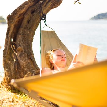 Beautiful girl lies on hammock on the beach reading book.