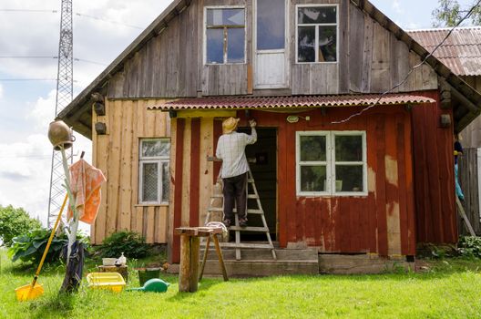 Painter farmer man with hat on ladder paint old wooden rural homestead house wall with brush paintbrush.