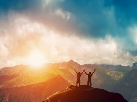 A happy couple standing together on the peak of a mountain with hands raised admiring breathtaking view at sunset