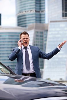 businessman shouting into the phone near the car and on the background of office