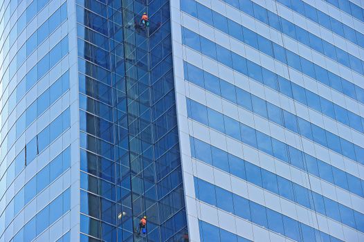 Workers suspended on a scaffold high up on a blue glass skyscraper
