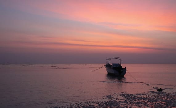 The low tide of predawn and dead calm sea immobilises this lone boat anchored near shoreline while the approaching dawn is reflected by the shades of blue and orange in the sky.