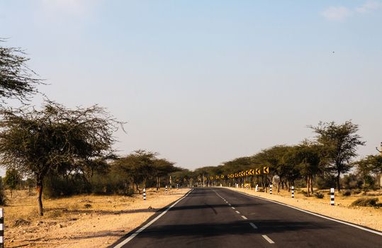 Asphalt covered road with a curve ahead to left and road signs is lined by trees on either side with almost cloudless blue sky providing relief on the way to sam sanddunes in jaisalmer, rajasthan, india.