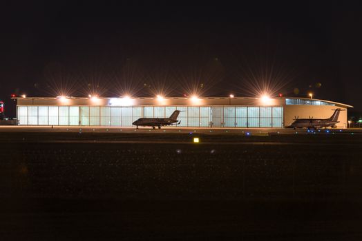 Two private jets parked in front of a lit hangar at dusk