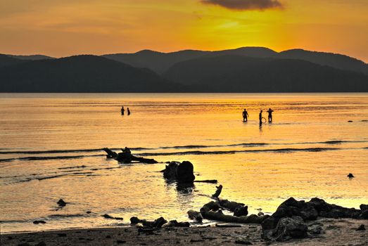 Orange dusk sky reflected in the sea with anonymous people frolicking and mountains in background.
