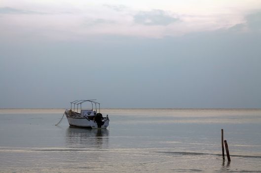 Grey sky reflected in the sea with ripples showing reflections of single boat anchored to wooden posts