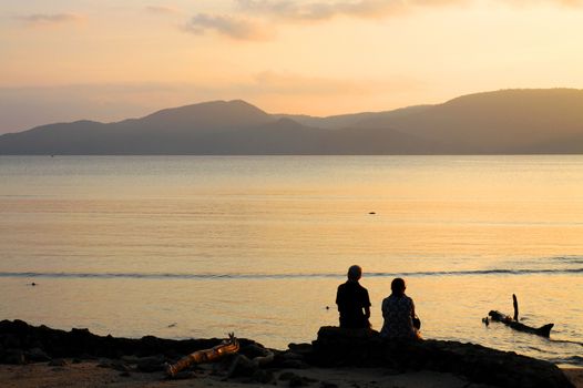 Silhouette  of a couple sitting on the Beach Twilight after  Sunset behind Mountains with calm seas reflecting the orange color of sky.