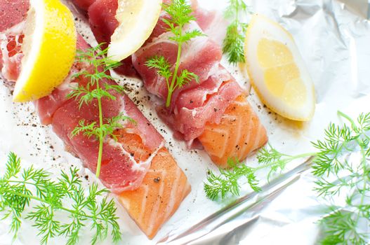 Fresh salmon fillets wrapped in bacon with lemon wedges and dill on kitchen foil