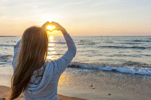 Blonde young girl holding hands in heart shape framing setting sun at sunset on ocean beach