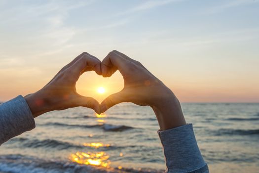 Hands and fingers in heart shape framing setting sun at sunset over ocean