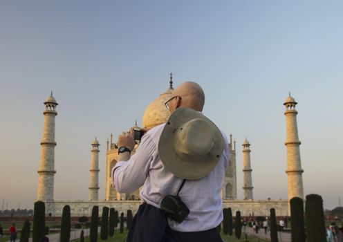 Waistup rearview of bald white adult male in casualwear with hat hanging from neck on back photographing taj mahal in agra india in golden yellow evening light