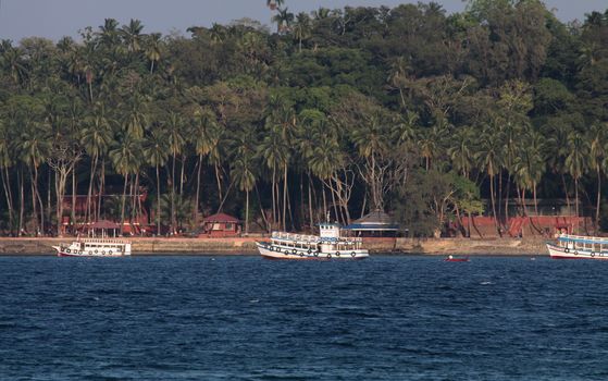 Dark Blue waters ,Seashore with a rowboat and three white boats at anchor and shore lined with coconut trees and Brown Buildings. Ross Island off Port Blair, Andamans, India.