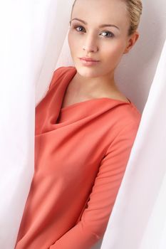 Attractive blonde woman in apricot dress looks from behind a white curtain.