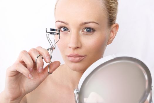 Portrait of a beautiful blonde with a curler for eyelashes and a mirror on a white background.