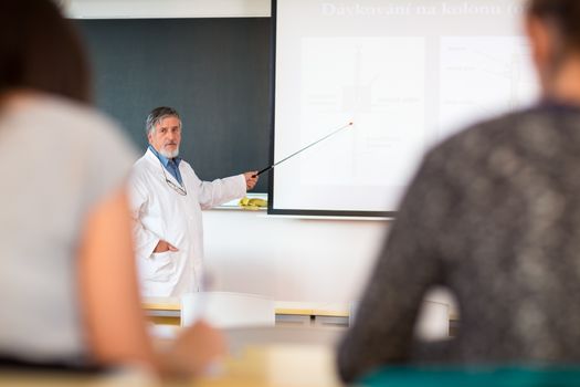 Senior chemistry professor giving a lecture in front of classroom full of students (shallow DOF; color toned image)