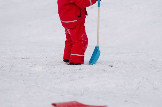 child stand with large snow shovel dressed in red waterproof clothing outdoor in winter time