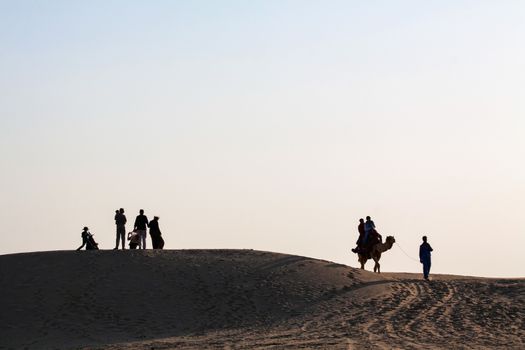 Silhouetted Family Group with Children watching sunset. Couple on Camel being led away by handler. sand dunes  with clear blue evening sky