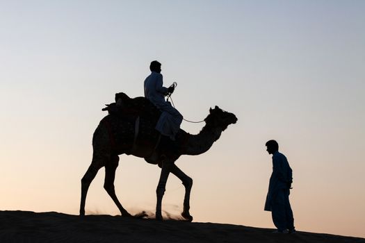 silhouetted against the bright orange hues of a dusk twilight, a single camel with rider is seen trotting away  past a male standing on the way side.