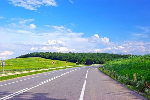 Summer road with road marking, meadow, forest and sky.
