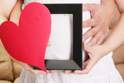 hands embracing a pregnant woman belly with her hands holding a wooden frame and a heart