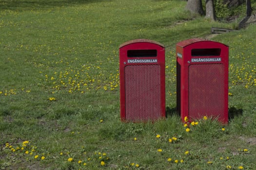 Two red garbage bins with text 'engangsgrillar' for disposable barbecue items in a park, Stockholm, Sweden in May.