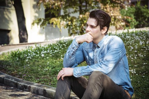 Young man sitting outdoors in public park, with hand on his chin looking away thinking