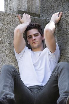 Attractive young man sitting on the ground against stone blocks outdoors, looking at camera, arms up