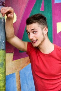 Attractive young blond man against colorful graffiti wall doing fun expression sticking out tongue