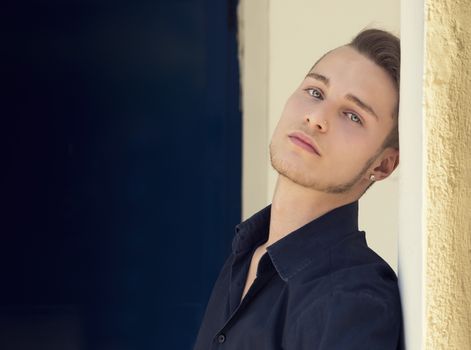 Attractive young blond man leaning against wall, looking at camera