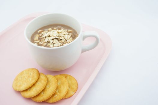 Cup of coco, oats and cracker placed on pink tray with white background.