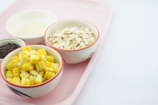 Corn, oats, chocolate and sweetened condensed milk placed on pink tray with white background.