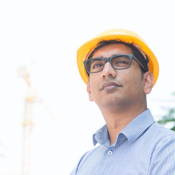 Portrait of an Asian Indian engineer looking away, standing in front construction crane, inspecting the progress of project.