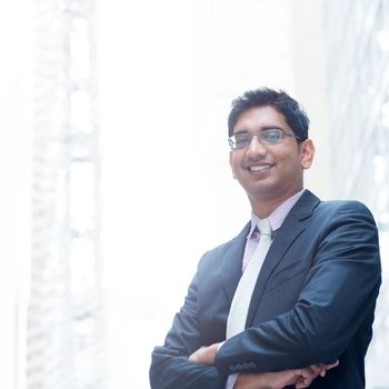 Portrait of a good looking smiling Indian businessman crossed arms standing at modern building, with natural light.
