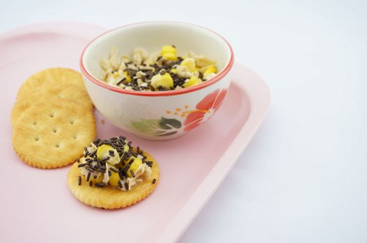 Mix corn, oats and sweetened condensed milk put on cracker sprinkle with chocolate placed on pink tray with white background.