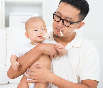 Asian family at home. Bad father smoking with baby together. Cigarette with lit and smoke. Unhealthy lifestyle or stop smoking concept photo.