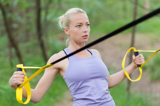 Young attractive woman does suspension training with fitness straps outdoors in the nature.