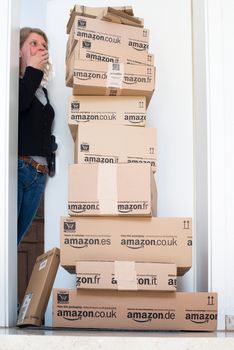 OSTFILDERN-SCHARNHAUSEN, GERMANY - MAY 18, 2014: A woman is horrified by a large stack of parcels by Amazon.com in different sizes waiting in front of the entrance door to her flat on May, 18, 2014 in Ostfildern-Scharnhausen near Stuttgart, Germany. This conceptual photo can serve different purposes: It might demonstrate the domination of Amazon.com in the area of online shopping or the trend in general to shop online for all the different items you need in your personal life.