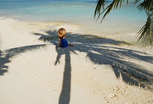 Woman in blue dress on a tropical beach at Maldives sitting in palm tree shadow