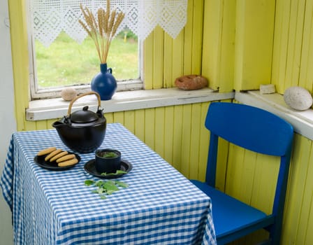 black ceramic tea set with mint tea cup and cookies on table with blue wooden chair in rural room