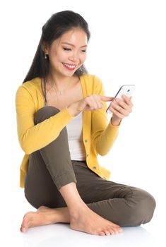 Full body Asian girl in yellow blouse texting with smartphone, seated on floor isolated on white background.