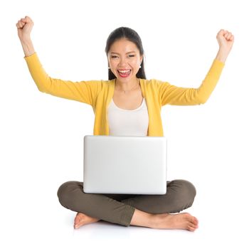 Full body Asian woman in yellow blouse arms up excited when using laptop computer notebook, seated on floor isolated on white background.