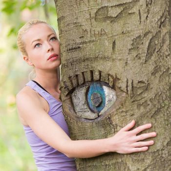 Relaxed young lady embracing a tree receiving life energy from the nature. Eye carved in tree trunk.