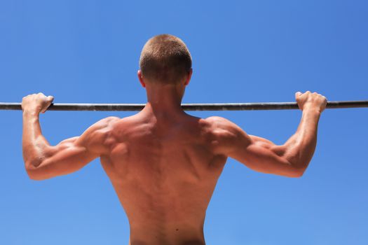 Young strong athlete doing exercise on horizontal bar against blue sky