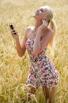 Young attractive woman is standing in a wheat field and listening to music with her smartphone. She looks happy and relaxed.