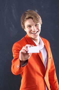 Portrait of a young, handsome man in the orange jacket standing on black background plate holding a business card with space for your text