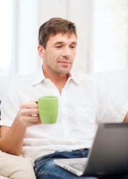 technology, drinks and lifestyle concept - man working with laptop at home, holding a cup of warm tea or coffee