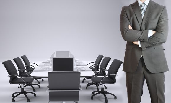 Businessman standing with his arms crossed. Conference table, chairs and laptops as backdrop