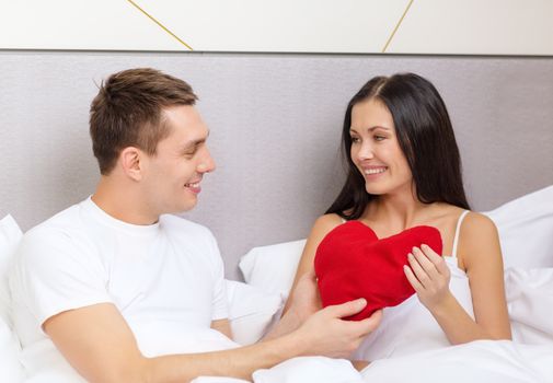 hotel, travel, relationships, holidays and happiness concept - smiling couple in bed with red heart-shaped pillow