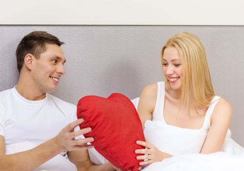 hotel, travel, relationships, holidays and happiness concept - smiling couple in bed with red heart-shaped pillow